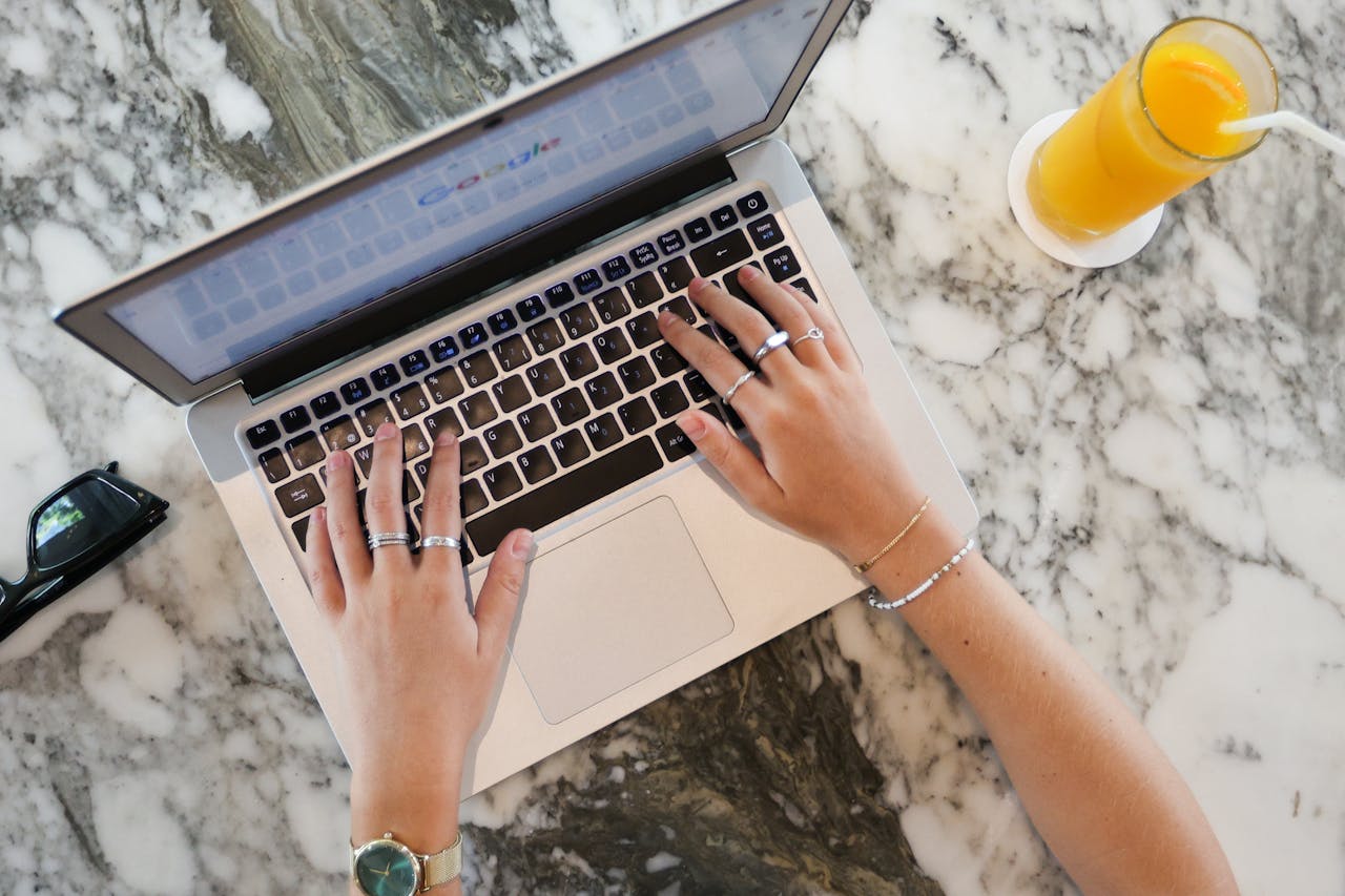 Services-01 Hands typing on a laptop with orange juice on a marble surface, ideal for remote work themes.